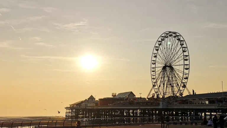 A sunset view of Blackpool’s Central Pier with the Ferris wheel silhouetted against a golden sky, people walking along the promenade, and birds flying overhead.