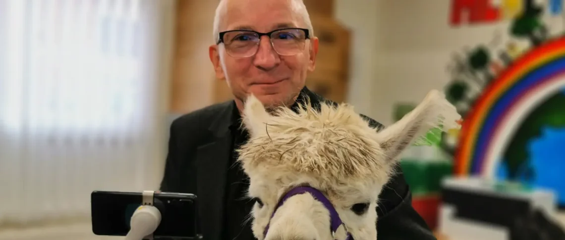 A man smiling while holding a white alpaca with a purple halter in an indoor community space.