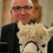 A man smiling while holding a white alpaca with a purple halter in an indoor community space.