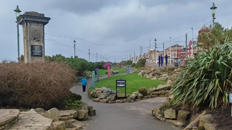 Entrance to Jubilee Gardens in Blackpool with a split pathway, decorative lamp posts, a 1925 commemorative plaque, lush planting, and a person walking with a cane.