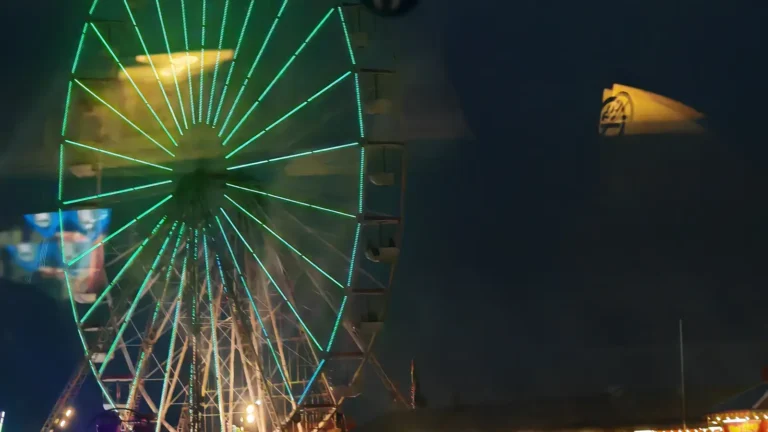 A night-time photograph of a large, brightly lit Ferris wheel in Blackpool. In the foreground, there are abstract, blurred reflections and streaks of light from an undefined source.