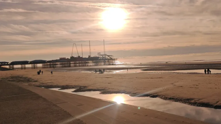Sunset in Blackpool. At the top of the photo, you can see the sun just above the horizon, with the end of the pier below it. The lower half of the frame shows the beach with a narrow strip of water after low tide, reflecting the sun and the pier.