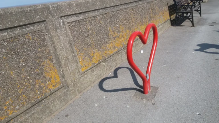 Heart‑shaped red bike rack casting a heart‑shaped shadow on a seaside promenade in Burnham‑on‑Sea.