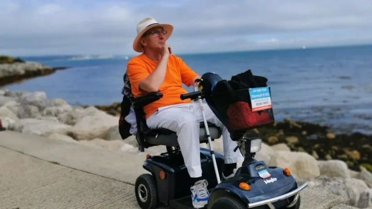The author sitting on a blue mobility scooter next to large stones arranged along a seaside sidewalk, with the sea visible in the background.