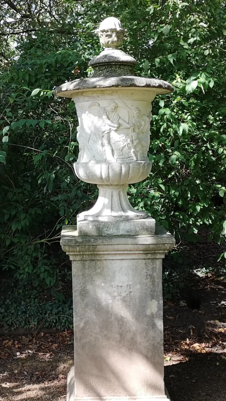 A classical stone urn at Shugborough Estate, decorated with relief figures and set on a pedestal among woodland.