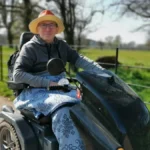 Close-up of the author sitting on a Tramper scooter, with his legs covered with a blanket and a blurred panorama in the background.