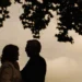 A couple standing in a tender embrace beneath a tree, silhouetted against a calm evening sky.