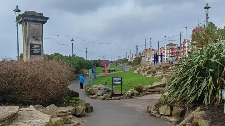Entrance to Jubilee Gardens in Blackpool with a split pathway, decorative lamp posts, a 1925 commemorative plaque, lush planting, and a person walking with a cane.