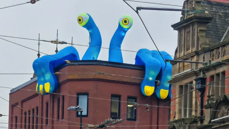 A large blue inflatable creature with bulging eyes and oversized yellow‑tipped limbs appears to climb over the roof of a brick building during the Blackpool Illuminations.