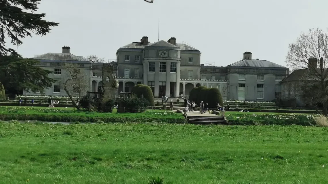 A riverside view of Shugborough Estate with its grand neoclassical façade, landscaped gardens and visitors walking near the entrance.