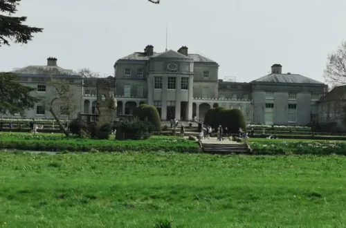 A riverside view of Shugborough Estate with its grand neoclassical façade, landscaped gardens and visitors walking near the entrance.