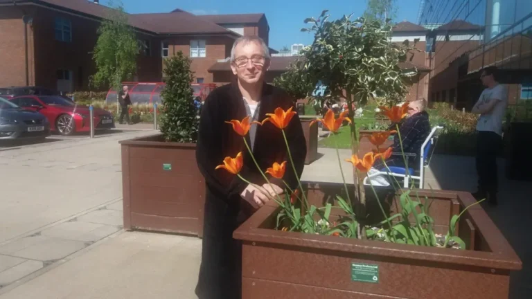 The author, wearing a dressing gown, stands in front of a hospital in Stoke-on-Trent, with a small flowerbed in front of him containing several orange tulips in bloom.