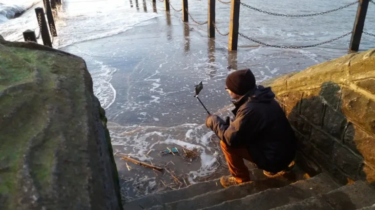 The blogger sits on the steps by the Marine Bay walkway in Weston‑super‑Mare, filming the sunset as waves rise against the stone.