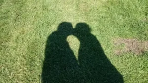 Shadows of two people on grass appearing to kiss, forming a heart‑like shape between their heads.