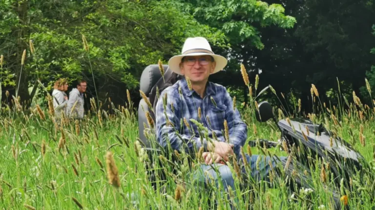 A man on a mobility scooter smiling among tall grass at Shugborough Estate, with two people walking in the background.