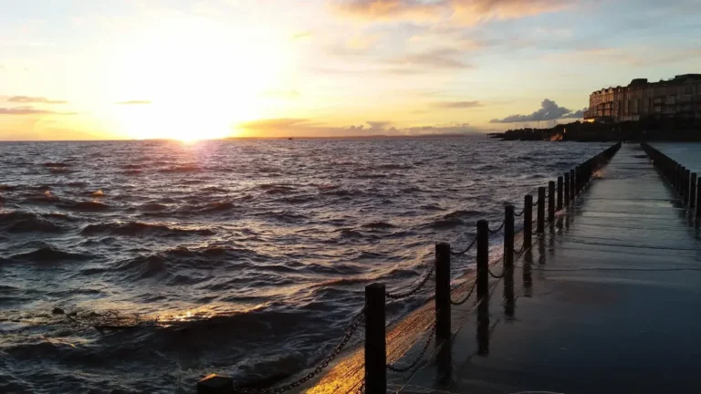 A romantic sunset view from the start of the walkway by Marine Bay in Weston‑super‑Mare, with rough waves glowing in the evening light and a distant building silhouetted on the shore.