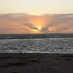 A warm sunset over the beach in Weston‑super‑Mare, with gentle waves and distant hills silhouetted on the horizon.