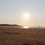A warm sunset over the beach in Weston‑super‑Mare, with people walking along the shoreline and distant hills silhouetted on the horizon.