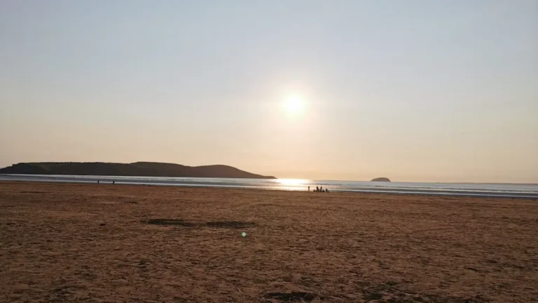 A warm sunset over the beach in Weston‑super‑Mare, with people walking along the shoreline and distant hills silhouetted on the horizon.
