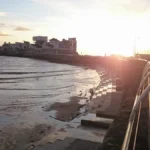 A sunset view from the promenade in Weston‑super‑Mare, looking toward Knightstone Island with a viewing telescope by the railings.