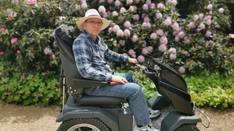 A side portrait shot of the author on a Tramper scooter, against a backdrop of blurred rhododendron bushes at Shugborough Estate.