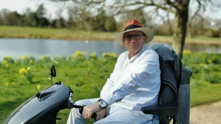 Close-up of the author sitting on a large mobility scooter, against a blurred background of yellow daffodils and the river at Shugborough Estate.