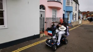 The author sitting on a mobility scooter against the backdrop of four houses with white, pink and blue plaster.