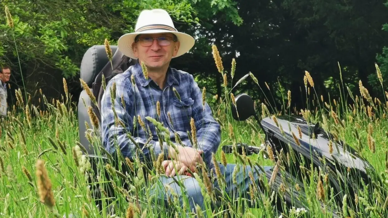 The author on a large scooter, the lower part of which is obscured by tall grass, wearing a colorful plaid shirt and a light-colored hat, at Shugborough Estate.