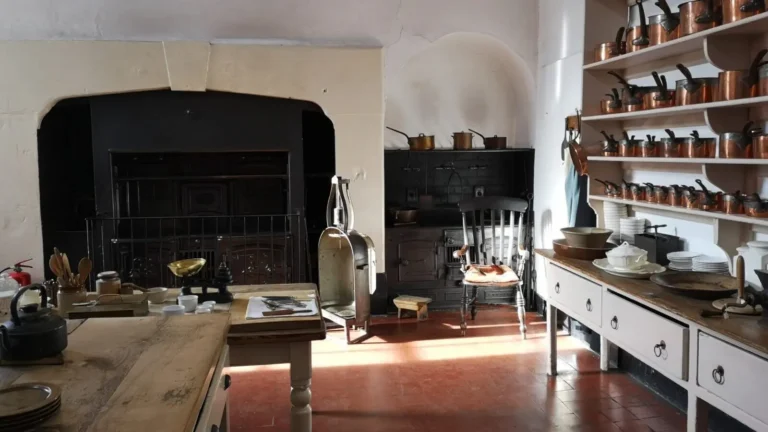 The interior of the main kitchen at Shugborough Estate, featuring a large fireplace, a stove, shelves with copper pots, and two work tables.