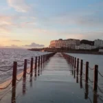 A wet walkway separating Marine Lake from the sea in Weston‑super‑Mare, with black posts and chains leading toward the town and hills.