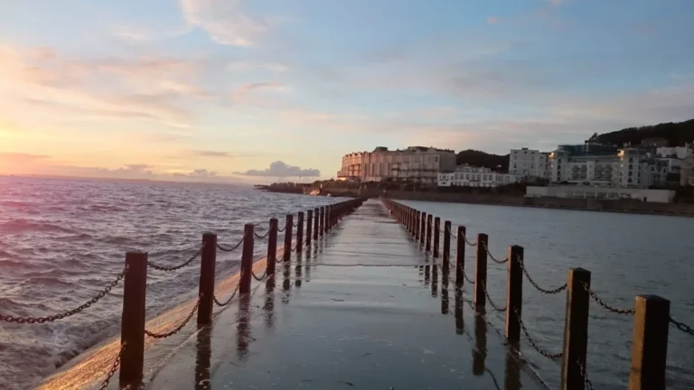 A wet walkway separating Marine Lake from the sea in Weston‑super‑Mare, with black posts and chains leading toward the town and hills.