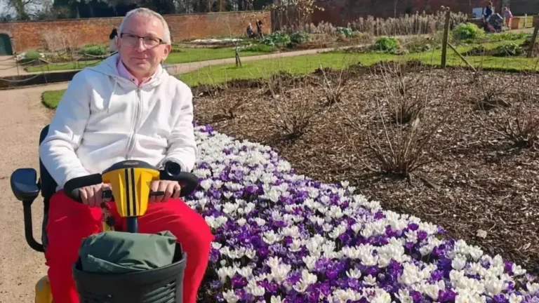 The author of the blog rests on his electric scooter on a paved path at Shugborough Estate, next to a long flowerbed with white and purple crocuses.