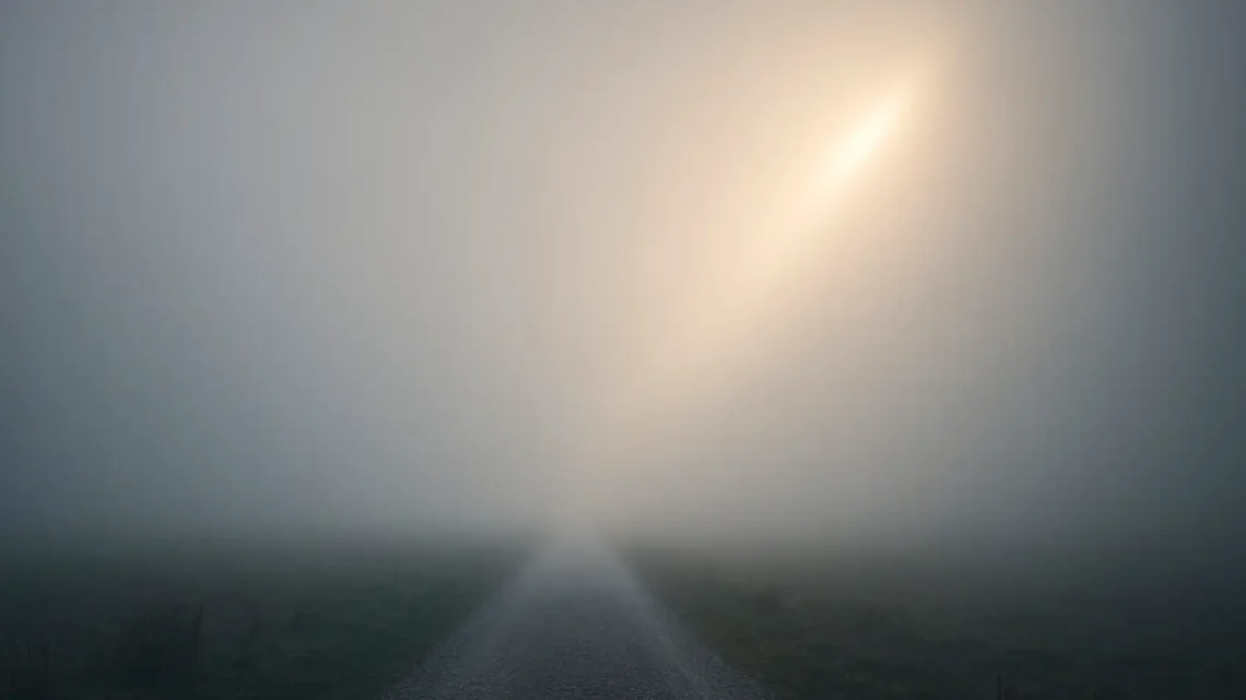 A foggy path stretching into the distance, with a ray of light breaking through — a quiet image of uncertainty and hope.