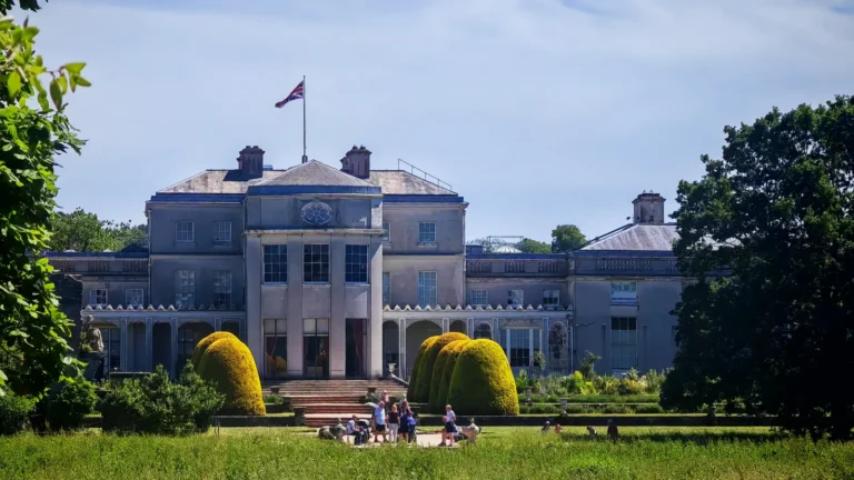 The Mansion at Shugborough Estate viewed from across the river, featuring classical architecture and terraced gardens with yellow bushes.